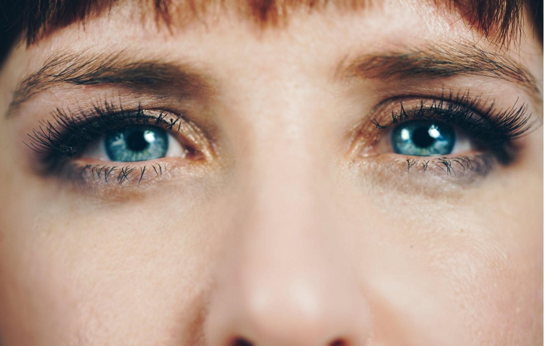 A woman looking into a camera with bluish-green eyes and long dark eye lashes