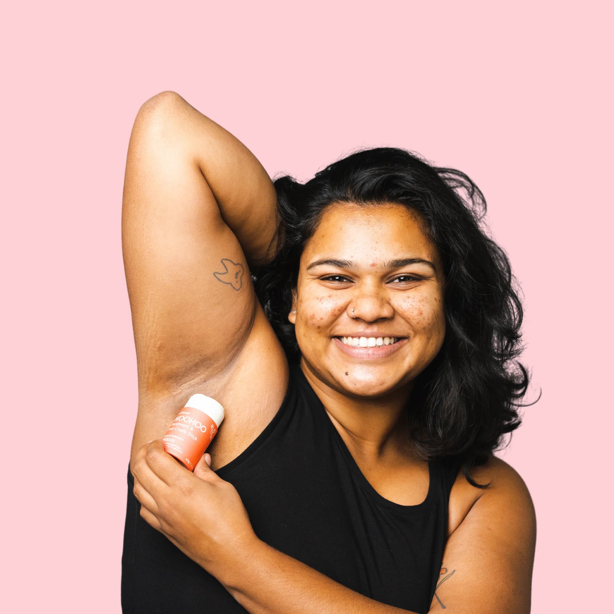Image of a woman in a black sleeveless shirt smiling to the camera and holding her arm up as she is applying an opened Urban Natural Deodorant Stick with her other hand in front of a pink background