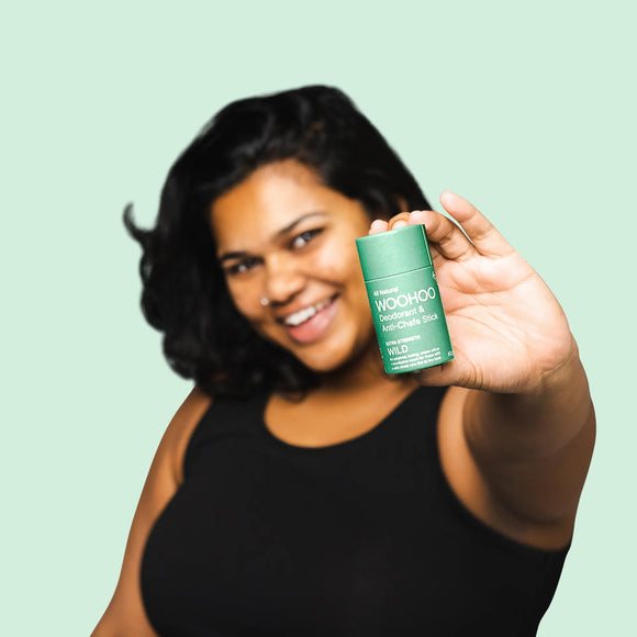 Image of a woman in a black sleeveless shirt smiling to the camera as she extends one hand with the Wild Natural Deodorant Stick towards the camera in front of a light green background