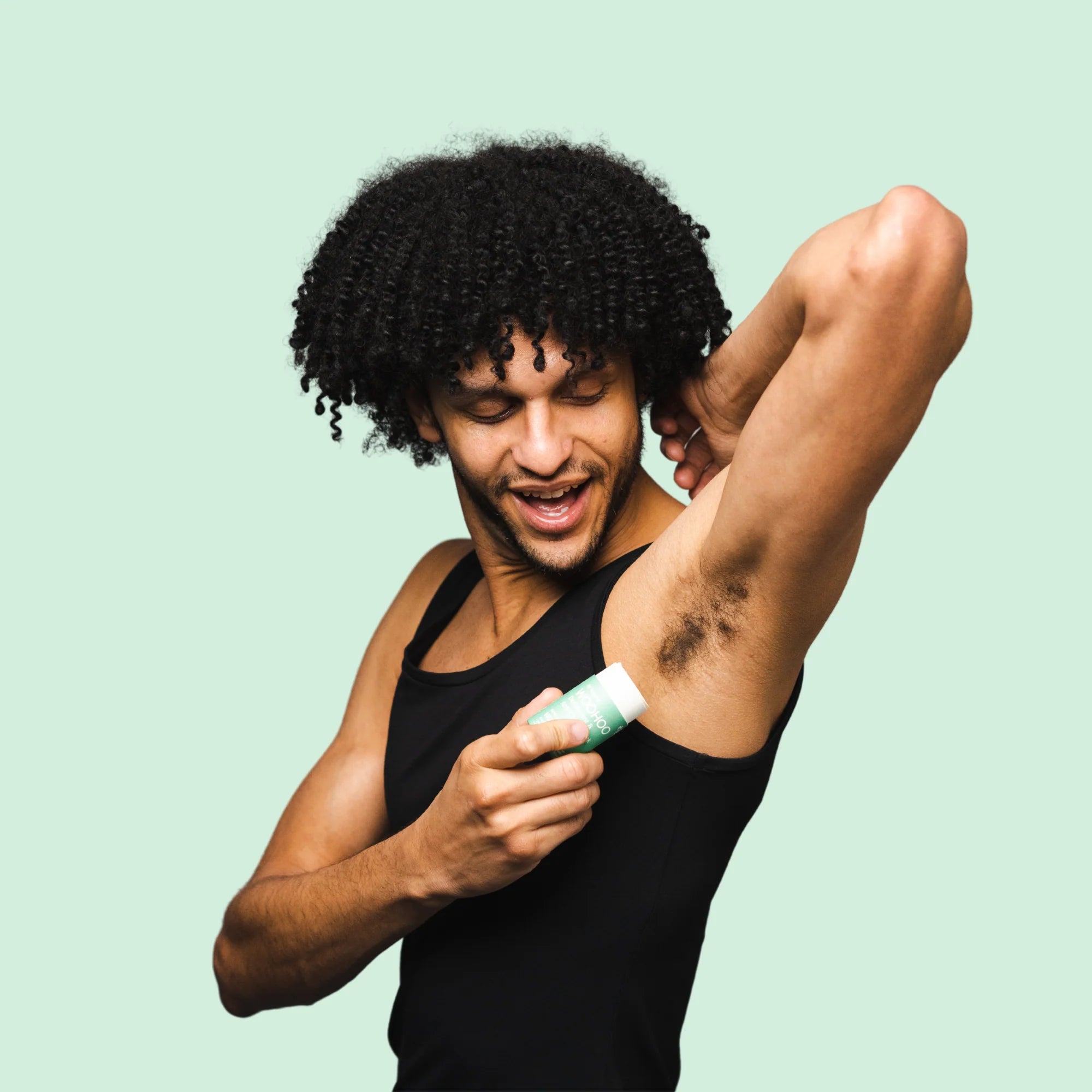 Image of a guy in a black sleeveless shirt holding his arm up as he is about to apply an opened Wild Natural Deodorant Stick with his other hand in front of a light green background